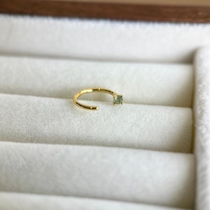 May include: A small, gold-coloured nose ring with a small, square, light green gemstone. The ring is open-ended and sits on a white, textured surface.