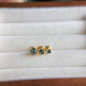 May include: Three gold stud earrings with round, moss-green gemstones of varying sizes. The earrings are displayed in a white, fabric-lined jewelry box. The gold settings provide a contrast to the green stones.