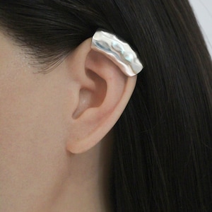 May include: A silver ear cuff with a hammered texture is shown on an ear. The jewellery piece is a curved bar that wraps around the ear's cartilage. The cuff is a bright, shiny silver colour.