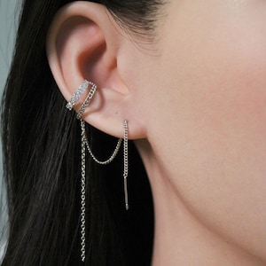 May include: Silver ear cuff and chain earrings. The ear cuff has a row of small, sparkling stones. A delicate chain dangles from the ear cuff, connecting to a chain earring that hangs down. The jewelry is worn on the ear.