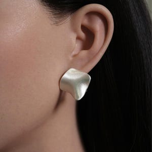 May include: A close-up of a silver-colored, abstract-shaped earring. The earring is a single stud, with a matte finish and a slightly curved, organic form. The earring is worn on an ear, with the skin visible in the background.