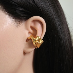 Puede incluir: Un ear cuff dorado con un diseño texturizado y orgánico. El cuff envuelve la oreja, presentando una forma escultural y fluida. La joya se lleva en la oreja, mostrando su forma artística única. El cuff es una pieza de joyería llamativa.