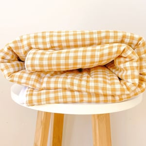 Peut inclure: Une couverture roulée, à carreaux jaunes et blancs, repose sur une petite table ronde blanche avec des pieds en bois. La couverture est pliée sur elle-même, mettant en valeur le motif carré répétitif. L'esthétique générale est simple et épurée.