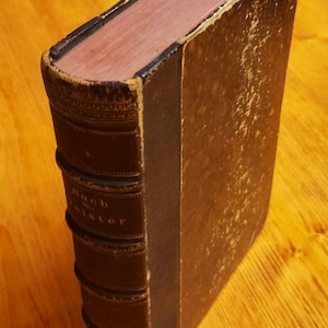 May include: An antique book with a brown leather cover, showing signs of age and wear. The spine has embossed text and decorative details. The pages have a reddish tint. The book's edges are worn, indicating its vintage.