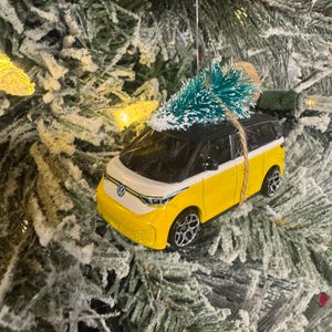 Puede incluir: Adorno navideño de coche de juguete amarillo y blanco con un pequeño árbol verde encima. El adorno cuelga de un árbol de Navidad cubierto de nieve. El coche tiene ruedas negras y techo negro.