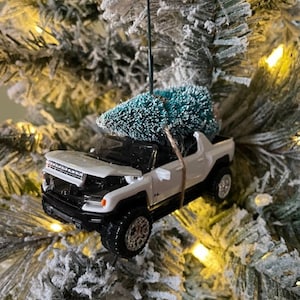 Puede incluir: Adorno de Navidad de camioneta blanca con un pequeño árbol verde en la parte superior. La camioneta está decorada con un lazo rojo y una campana plateada.