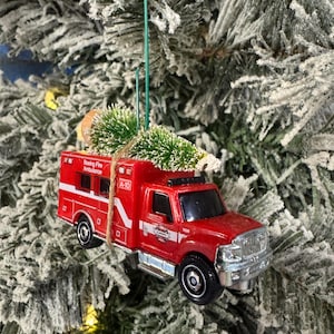 以下が含まれることがあります： 赤色の救急車のクリスマスオーナメント。銀色のアクセントと黒い車輪が付いています。「Boeing Fire Ambulance」と「A-10」の文字がプリントされています。小さな緑色のクリスマスツリーが上部に付いています。