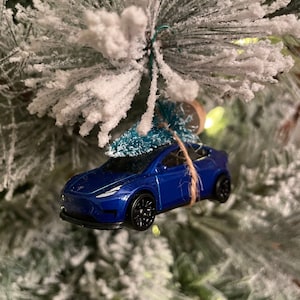 May include: A blue toy car Christmas ornament with black wheels hangs from a snowy Christmas tree branch. The ornament is suspended by twine and a small green tree. The car has a Tesla logo.
