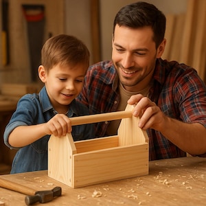 Puede incluir: Una caja de herramientas de madera de color claro con un asa es sostenida por un hombre y un niño pequeño. Un martillo y trozos de madera están sobre la mesa de madera. El hombre lleva una camisa a cuadros y el niño una camisa vaquera. La escena tiene lugar en un taller.