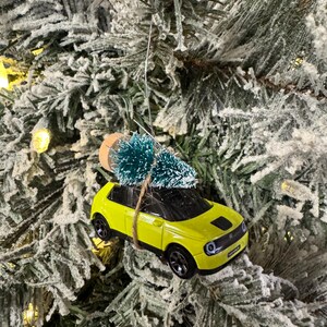 May include: A yellow toy car Christmas ornament with a small green tree tied to the roof. The ornament hangs from a silver wire against a backdrop of a snow-covered Christmas tree.
