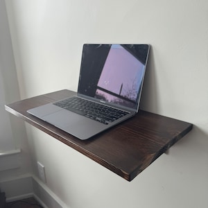 May include: A dark brown wooden floating shelf mounted to a white wall with a silver laptop computer on top of the shelf.