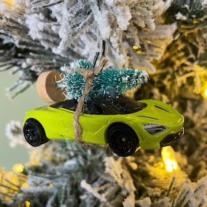 May include: A green toy car with a small green Christmas tree on top. The car is hanging from a Christmas tree branch.