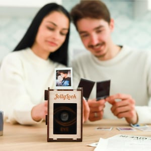Pu&ograve; includere: Una macchina fotografica in legno con le parole "JollyLook" e "EYE" stampate su di essa. La macchina fotografica &egrave; appoggiata su un tavolo con una coppia sullo sfondo.