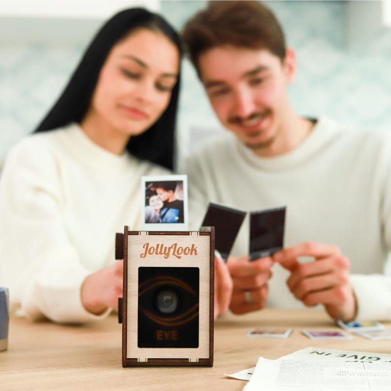 Pu&ograve; includere: Una macchina fotografica in legno con le parole "JollyLook" e "EYE" stampate su di essa. La macchina fotografica &egrave; appoggiata su un tavolo con una coppia sullo sfondo.