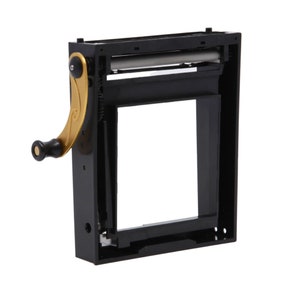 Op de afbeelding: Een zwarte metalen sigarettenrolmachine met een gouden draaiknop. De machine heeft een transparant venster om het rolproces te bekijken.