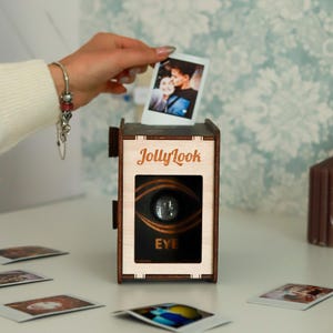 Pu&ograve; includere: Una scatola di legno a forma di macchina fotografica con uno slot per inserire le foto. La scatola ha un design a occhio bianco e nero con la parola "EYE" stampata sotto. La scatola &egrave; etichettata "JollyLook".