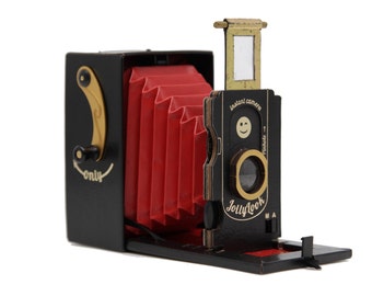 Puede incluir: Una cámara instantánea de estilo vintage negra y dorada con fuelle rojo. La cámara tiene una carita sonriente en la parte delantera y el texto "JollyLook" e "Instant camera".