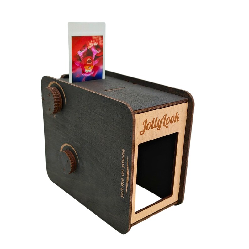 Pu&ograve; includere: Una scatola di legno nera a forma di macchina fotografica con una fessura per una foto. La scatola ha un ritaglio per un telefono e il testo "JollyLook" e "put me on phone" &egrave; stampato sul lato.