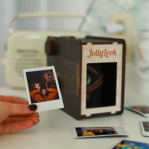 Pu&ograve; includere: Una macchina fotografica in legno marrone scuro con il testo "JollyLook" stampato sul davanti. Una foto Polaroid bianca con una persona al suo interno viene tenuta davanti alla macchina fotografica.