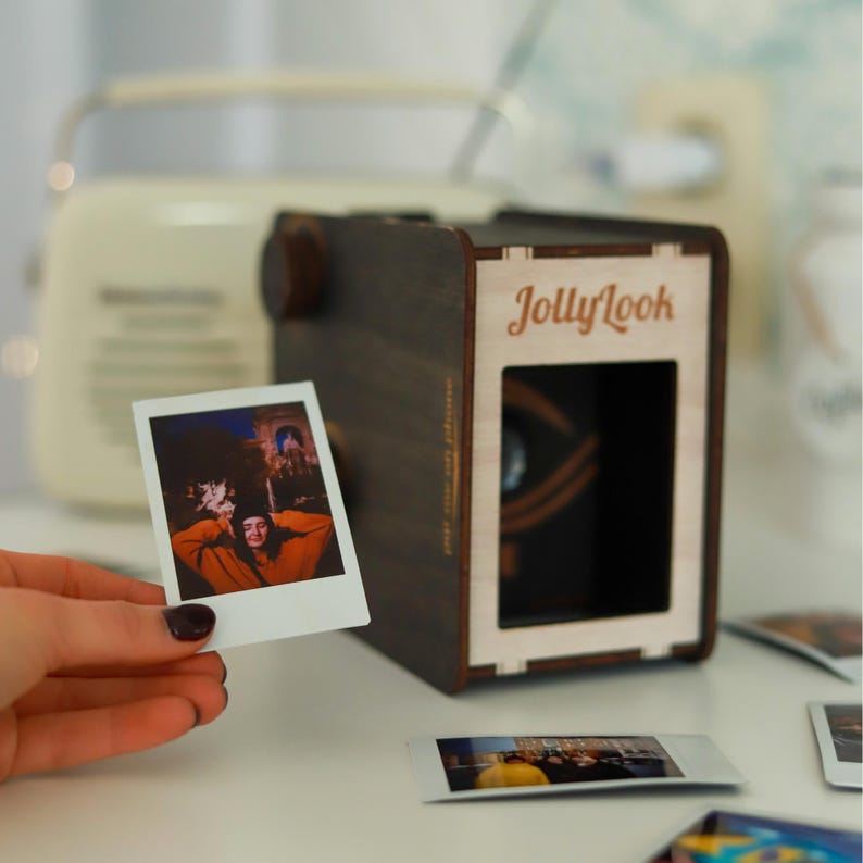 Pu&ograve; includere: Una macchina fotografica in legno marrone scuro con il testo "JollyLook" stampato sul davanti. Una foto Polaroid bianca con una persona al suo interno viene tenuta davanti alla macchina fotografica.
