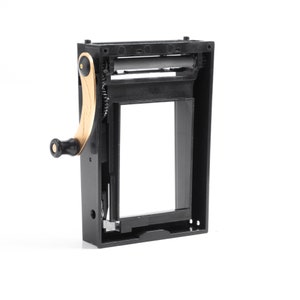 Könnte beinhalten: Schwarzer Plastik-Filmhalter mit einem goldfarbenen Kurbelgriff. Der Halter hat eine rechteckige Öffnung zum Einlegen von Film.