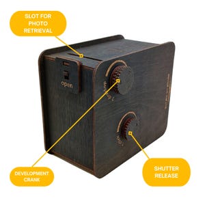 Pu&ograve; includere: Una scatola di legno scuro a forma di macchina fotografica con uno slot per il recupero delle foto, una manovella di sviluppo e un pulsante di scatto. La scatola ha un'etichetta che dice "Stampa le tue foto".