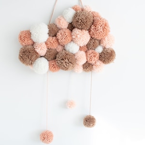 May include: A cloud-shaped wall hanging made of brown, white and pink yarn pom-poms. The pom-poms are strung together and hang from a string. Three smaller pom-poms hang from the bottom of the cloud.