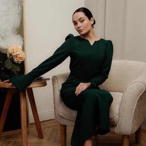 May include: A woman wearing a dark green dress with a v-neck and long sleeves. She is sitting on a light beige armchair with wooden legs. The woman is wearing black heels.