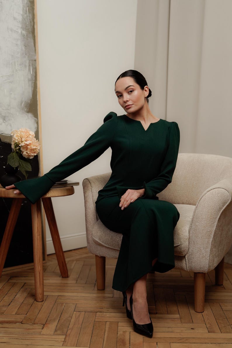 May include: A woman wearing a dark green dress with a v-neck and long sleeves. She is sitting on a light beige armchair with wooden legs. The woman is wearing black heels.