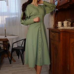 May include: A woman wearing a long-sleeved, sage green dress with a button-up front. The dress has a fitted bodice and a flowing skirt. She is wearing white heels and has her hair pulled back in a braid.