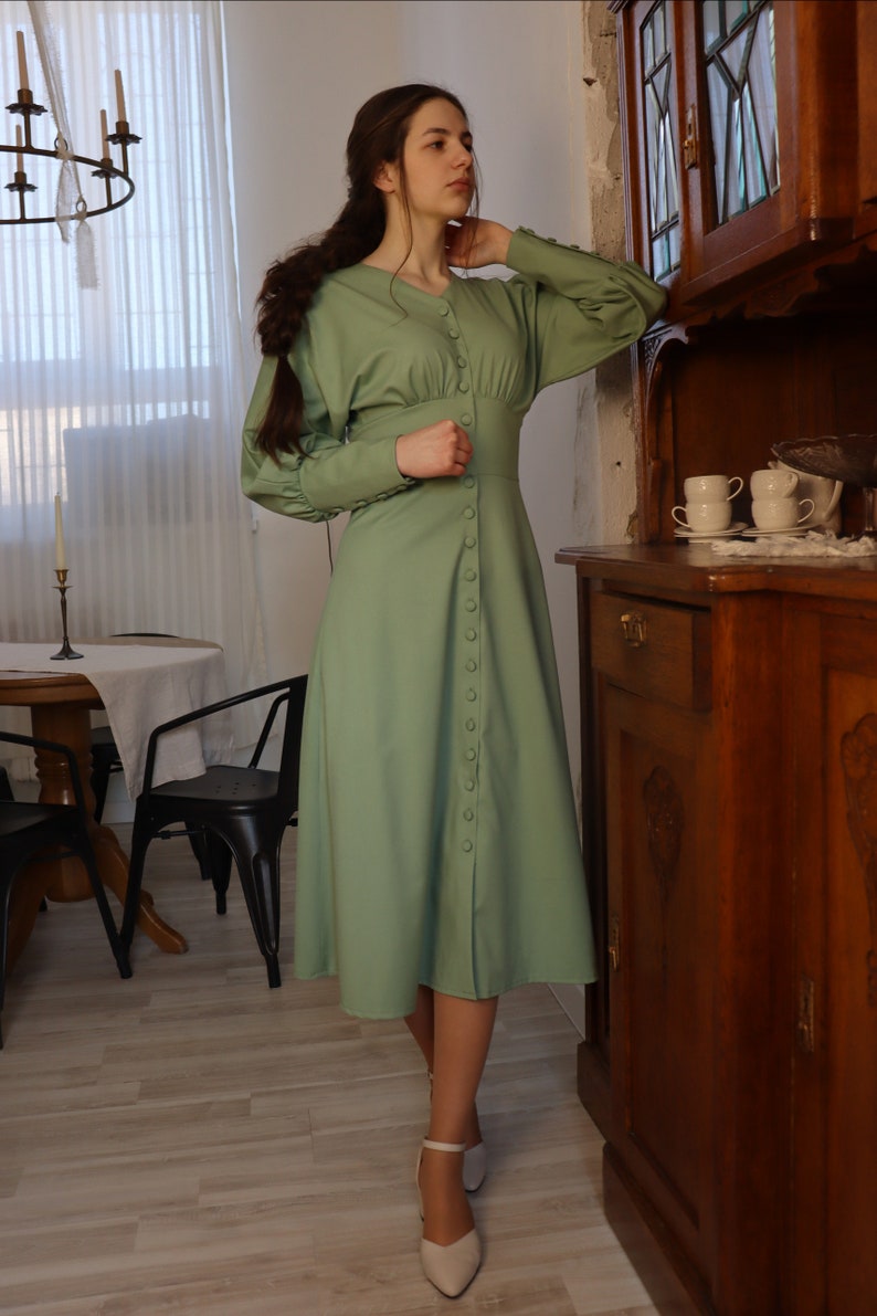 May include: A woman wearing a long-sleeved, sage green dress with a button-up front. The dress has a fitted bodice and a flowing skirt. She is wearing white heels and has her hair pulled back in a braid.