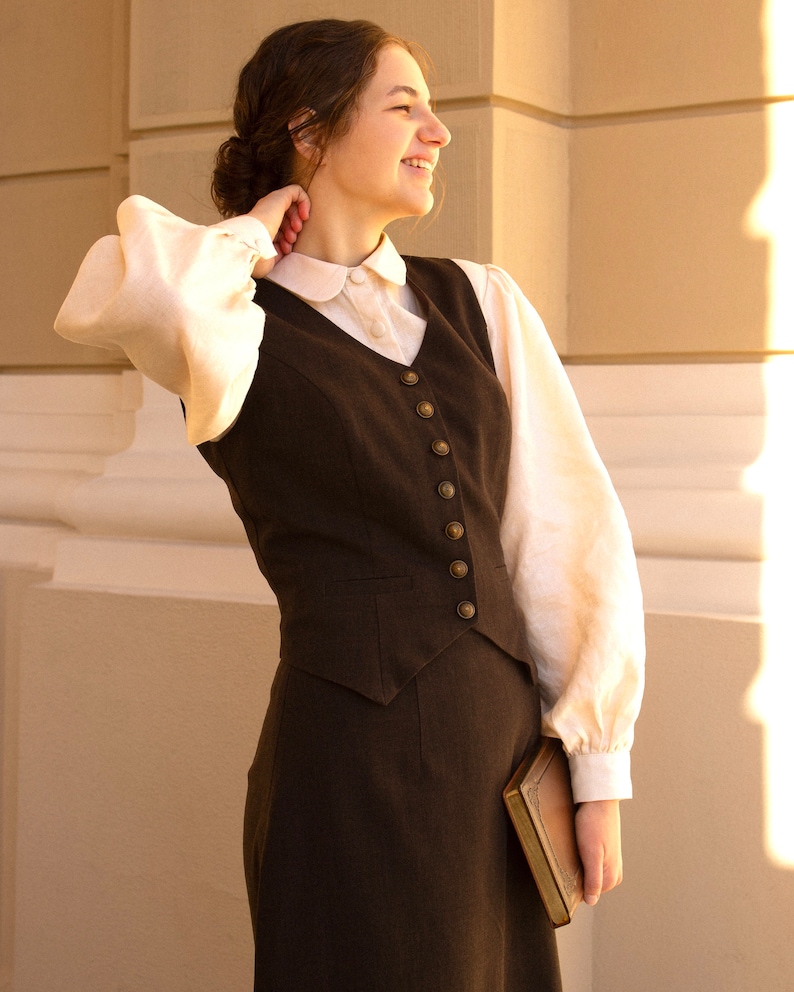 May include: A woman wearing a brown vest and skirt with a white blouse and a brown book in her hand.