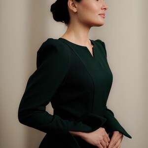 May include: A woman wearing a dark green dress with a V-neck and long sleeves. The dress has a fitted bodice and a slightly flared skirt.