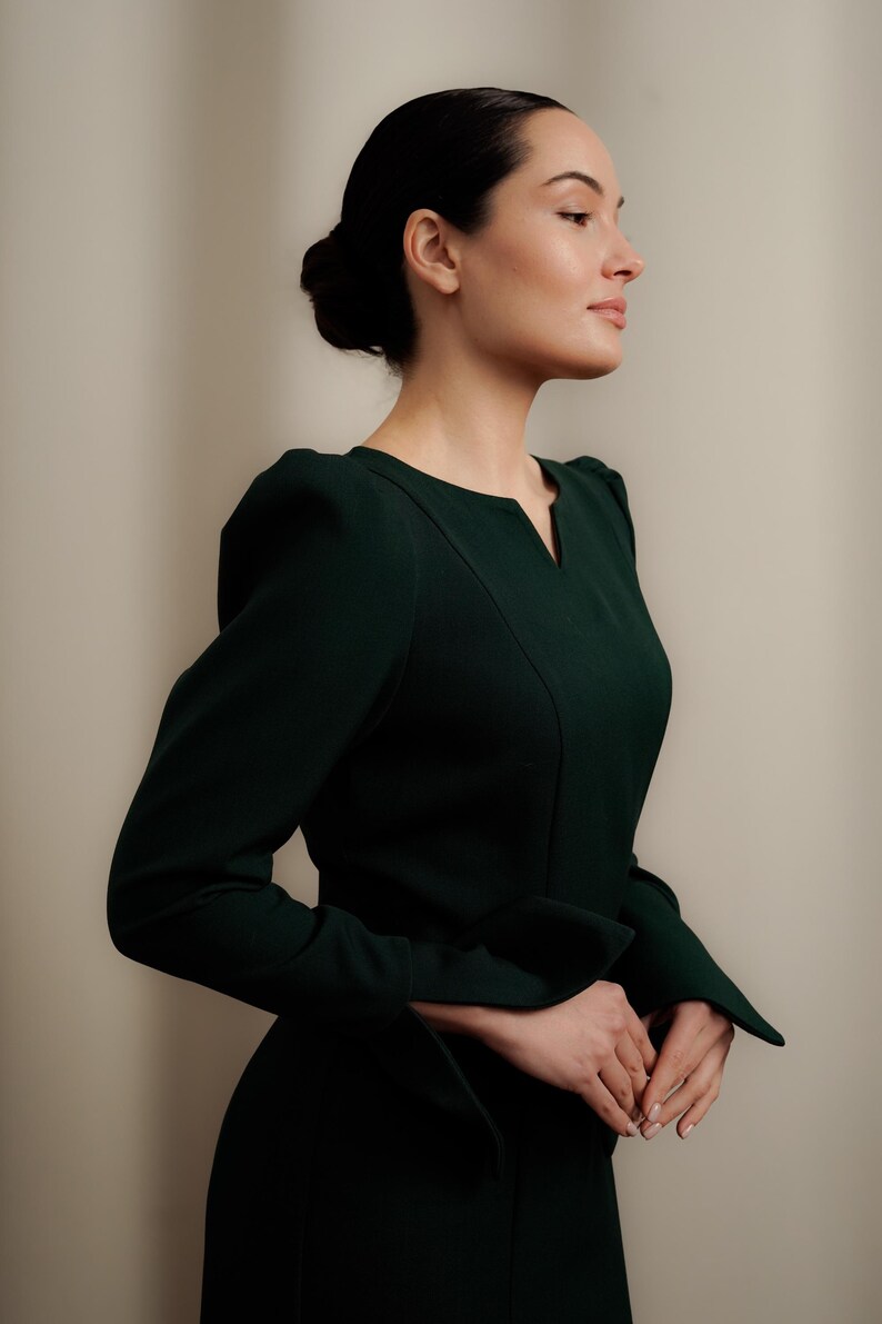 May include: A woman wearing a dark green dress with a V-neck and long sleeves. The dress has a fitted bodice and a slightly flared skirt.