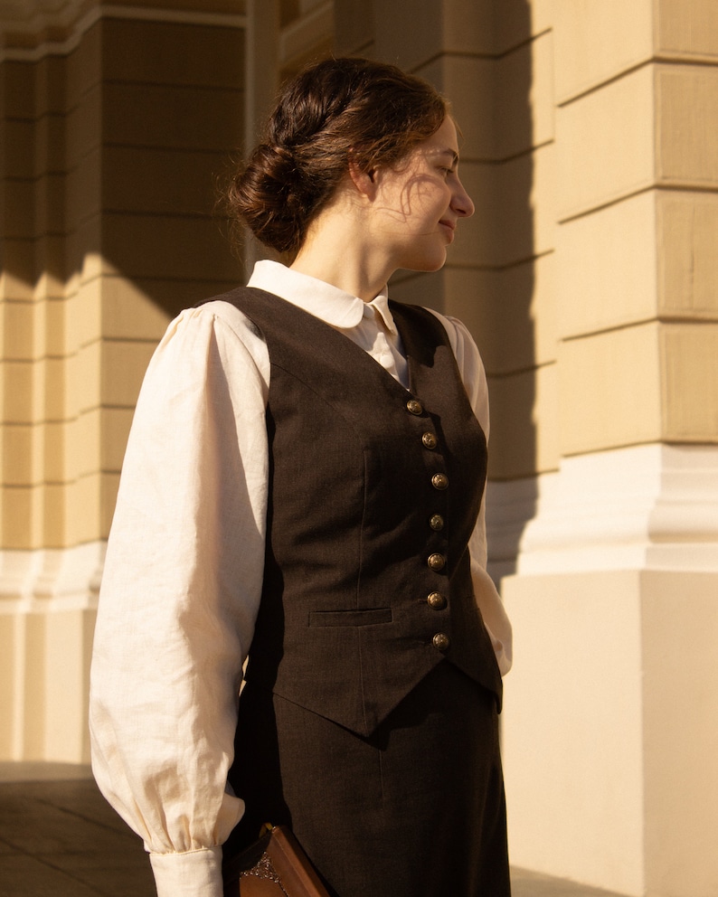 May include: A woman wearing a brown vest with gold buttons over a white blouse with a peter pan collar. She is standing in front of a building with a light brown facade.