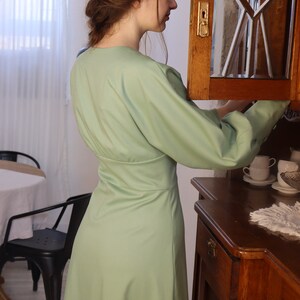 May include: A woman wearing a light green dress with long sleeves and a v-neck. She is standing in front of a wooden cabinet with a glass door.