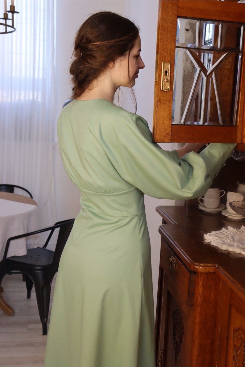 May include: A woman wearing a light green dress with long sleeves and a v-neck. She is standing in front of a wooden cabinet with a glass door.