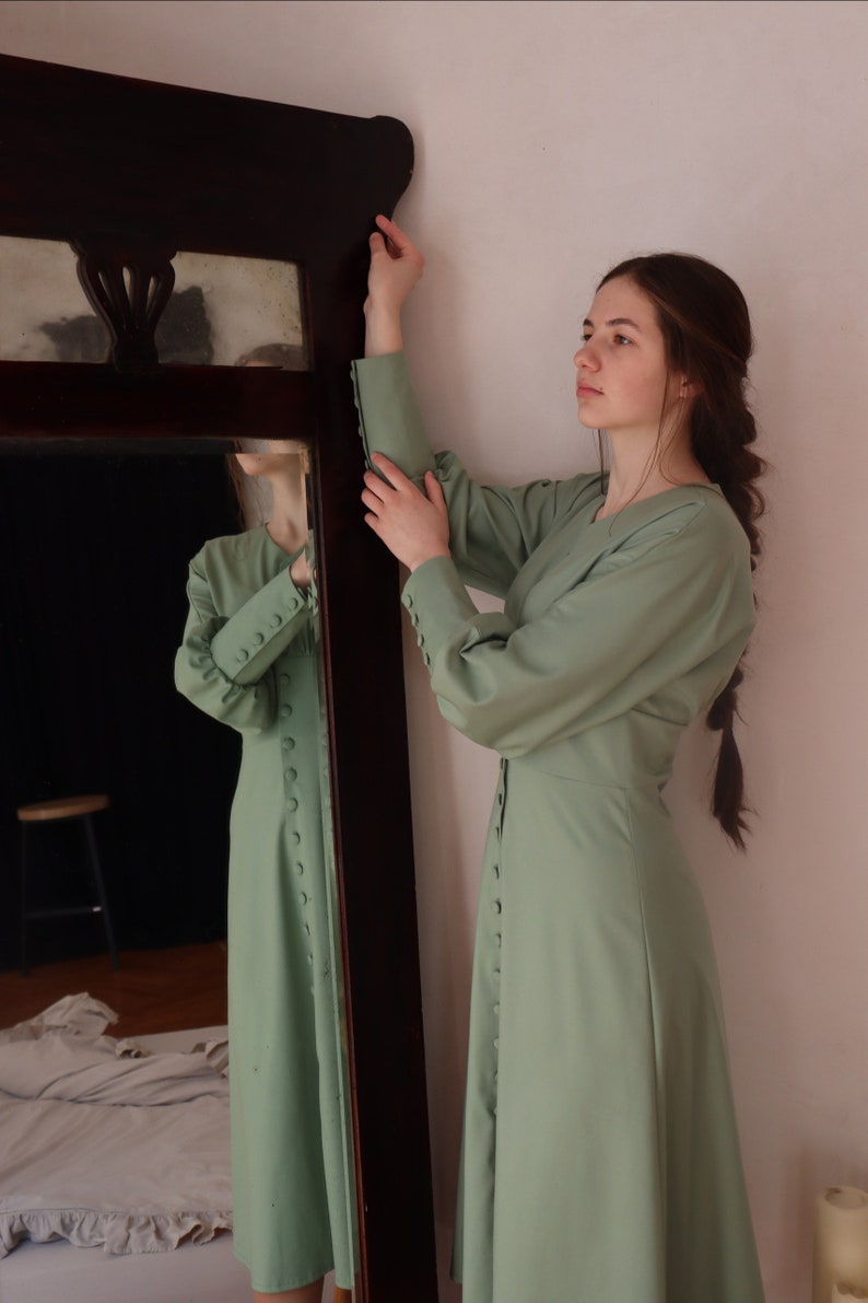 May include: A woman wearing a sage green dress with long sleeves and a button-up front stands in front of a mirror. The dress has a slightly flared skirt and the woman is looking to the side.