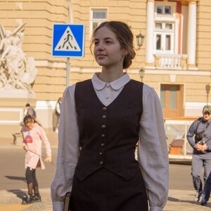 May include: A woman wearing a white collared shirt with a brown vest and a brown skirt. She is holding a book in her left hand and walking down a street.
