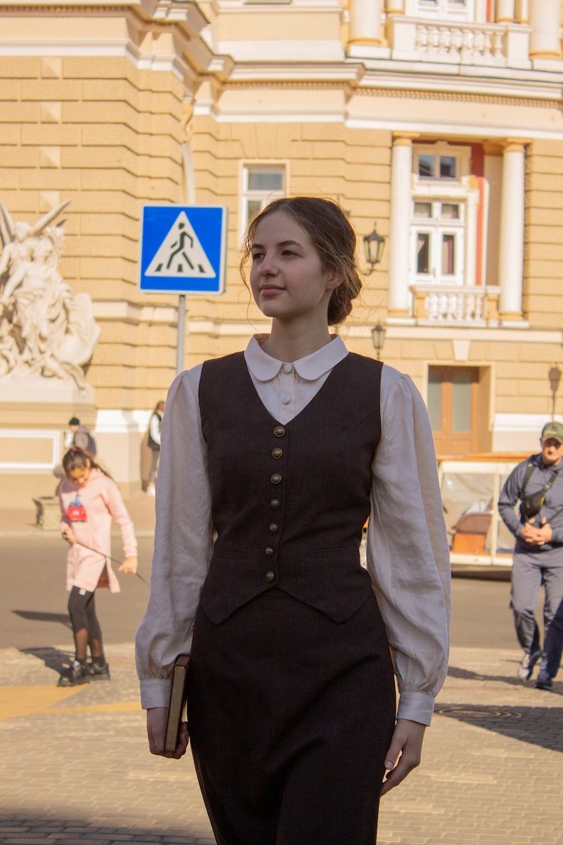 May include: A woman wearing a white collared shirt with a brown vest and a brown skirt. She is holding a book in her left hand and walking down a street.