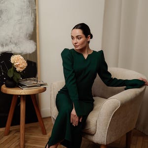 May include: A woman wearing a dark green dress with long sleeves and a V-neck sits in a light beige armchair. She is wearing black heels and has her hair pulled back.