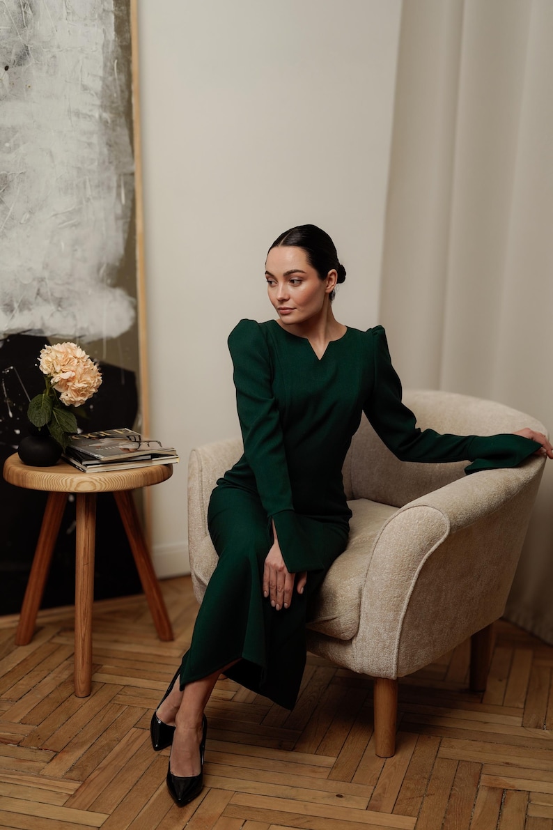 May include: A woman wearing a dark green dress with long sleeves and a V-neck sits in a light beige armchair. She is wearing black heels and has her hair pulled back.