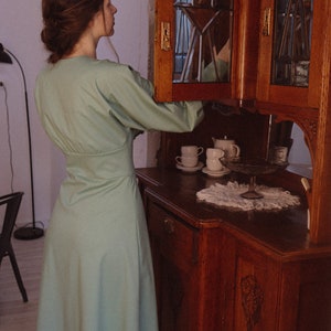 May include: A woman wearing a light green dress stands in front of a wooden cabinet with glass doors. The cabinet is open, revealing white teacups and a teapot.