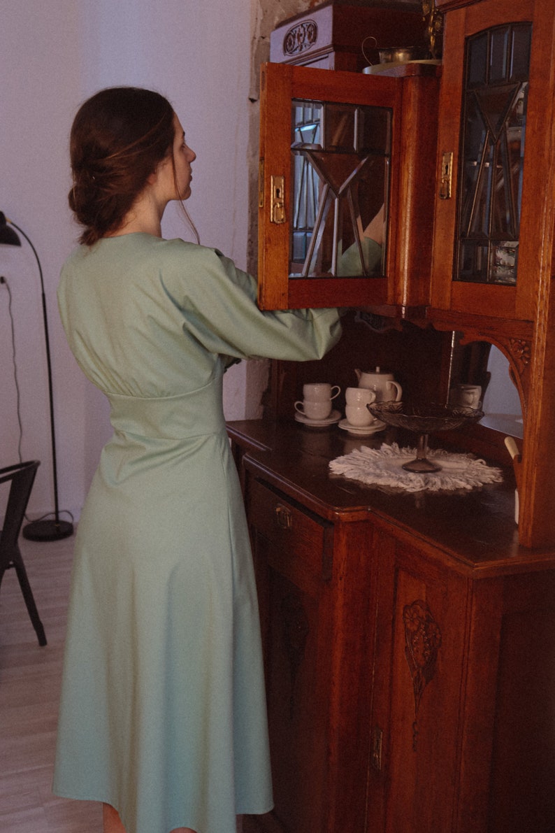 May include: A woman wearing a light green dress stands in front of a wooden cabinet with glass doors. The cabinet is open, revealing white teacups and a teapot.