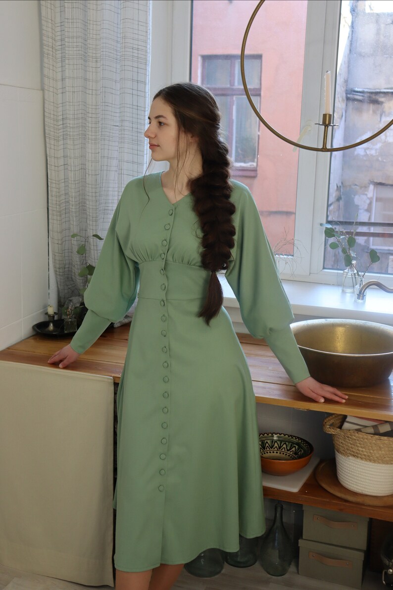 May include: A sage green midi dress with long sleeves and a button-up front. The dress has a fitted bodice and a flowy skirt.