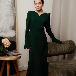 May include: A woman wearing a dark green long-sleeved dress with a v-neck and a slightly flared skirt. She is standing in front of a light-colored wall with a painting behind her.