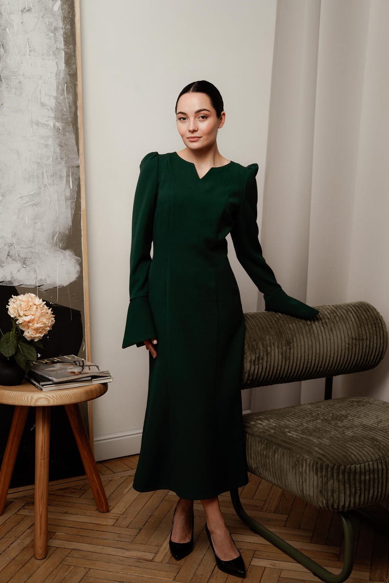 May include: A woman wearing a dark green long-sleeved dress with a v-neck and a slightly flared skirt. She is standing in front of a light-colored wall with a painting behind her.
