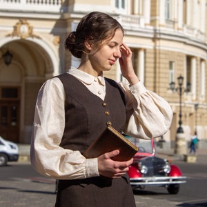 May include: A woman wearing a brown vest over a white blouse and a brown skirt, holding a brown book in front of a building.