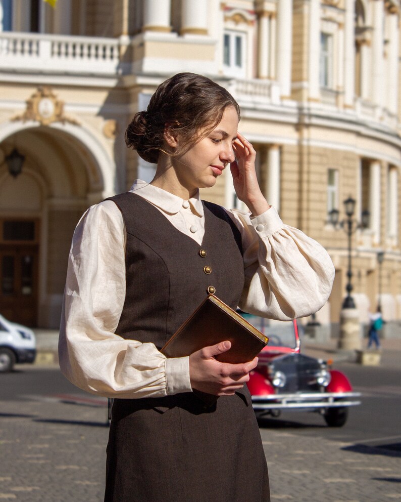 May include: A woman wearing a brown vest over a white blouse and a brown skirt, holding a brown book in front of a building.