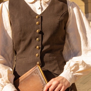 May include: A brown vest with gold buttons worn over a white collared shirt with puffed sleeves. The person is holding a brown leather-bound journal.